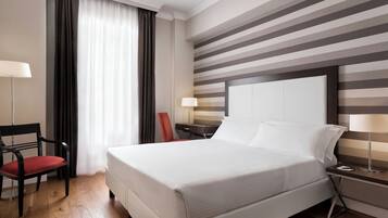 A hotel room with a large bed, a desk, a chair, a lamp, and a window with curtains.