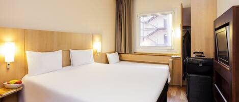 Habitación de hotel con una cama grande, lámparas de noche, una mesita con frutero, una maleta y un televisor.