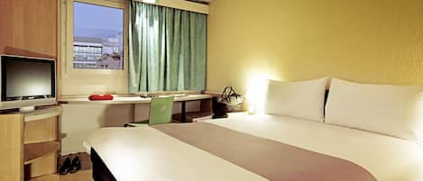 Una habitación de hotel con una cama, un escritorio con televisor, una silla, una ventana con cortinas y vistas a edificios.