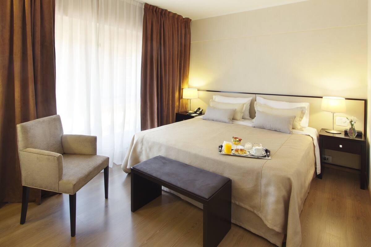 Hotellihuone, jossa on suuri sänky, tuoli, penkki, yöpöytä lampun kanssa ja toinen pienempi pöytä sängyn vieressä, jossa on myös lamppu.