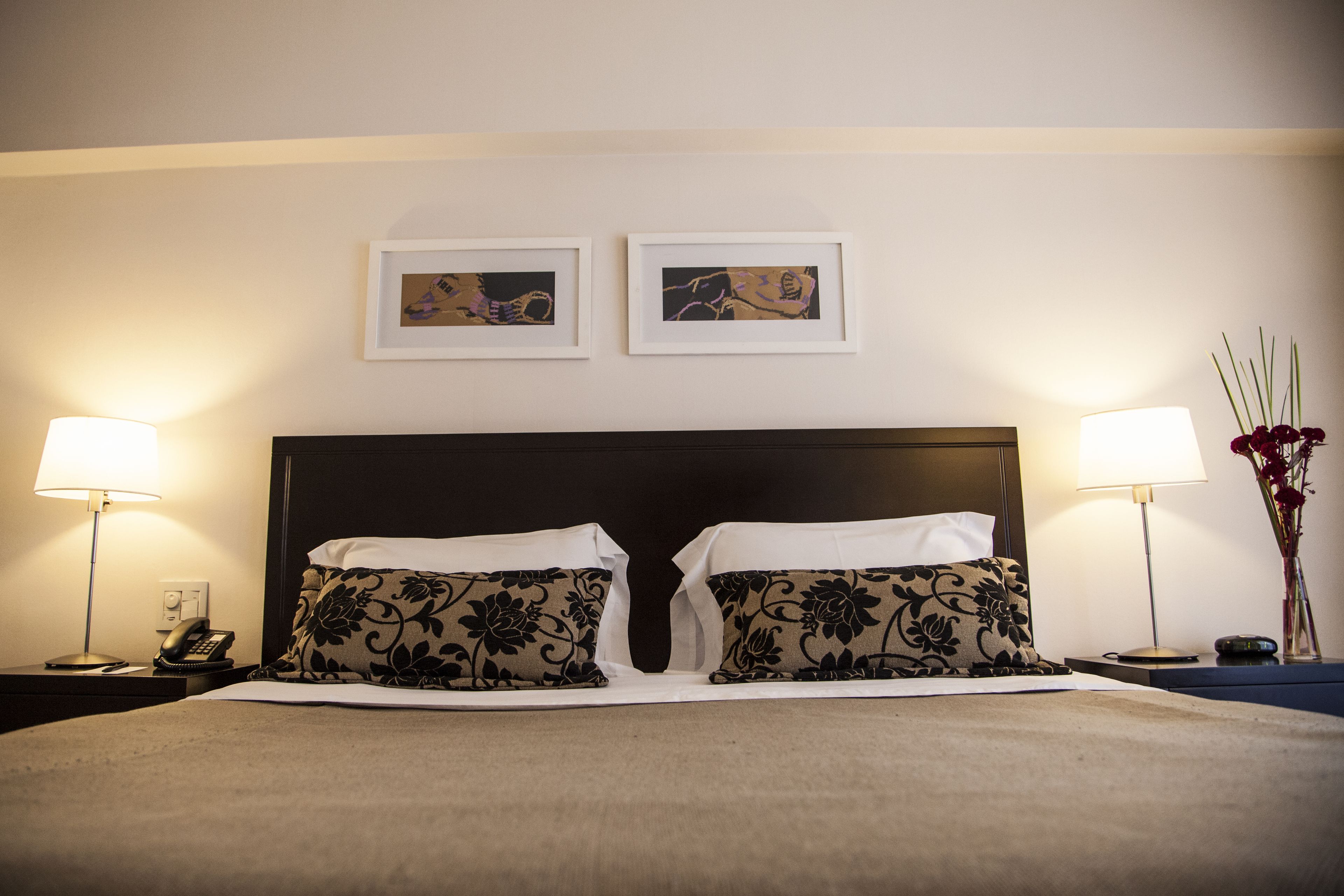A neatly made bed with floral-patterned pillows, two framed artworks above it, and bedside lamps on either side.