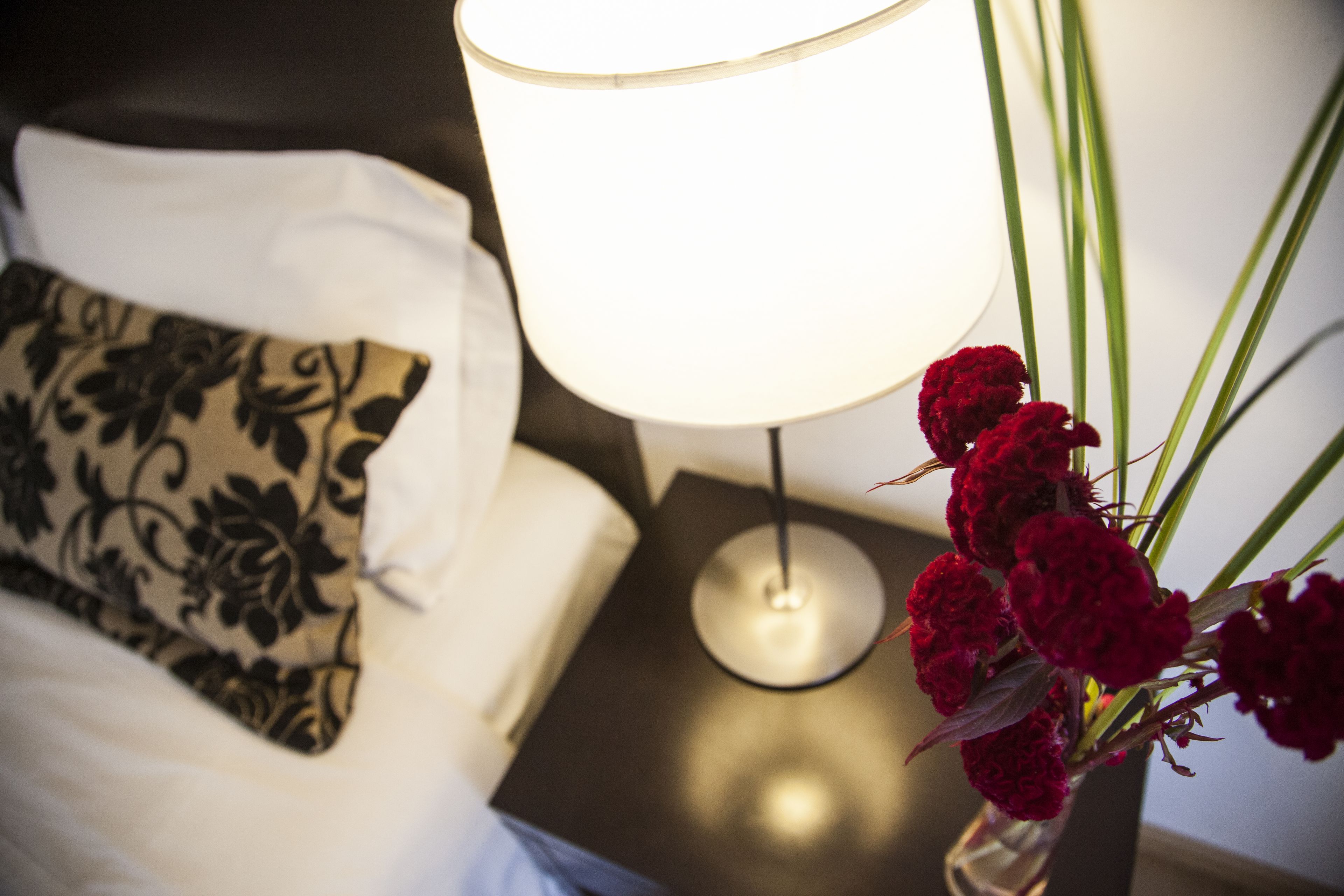 Ein Bett mit einem schwarz-weiß gemusterten Kopfkissen, ein Nachttisch mit einer Lampe und eine Vase mit roten Blumen.