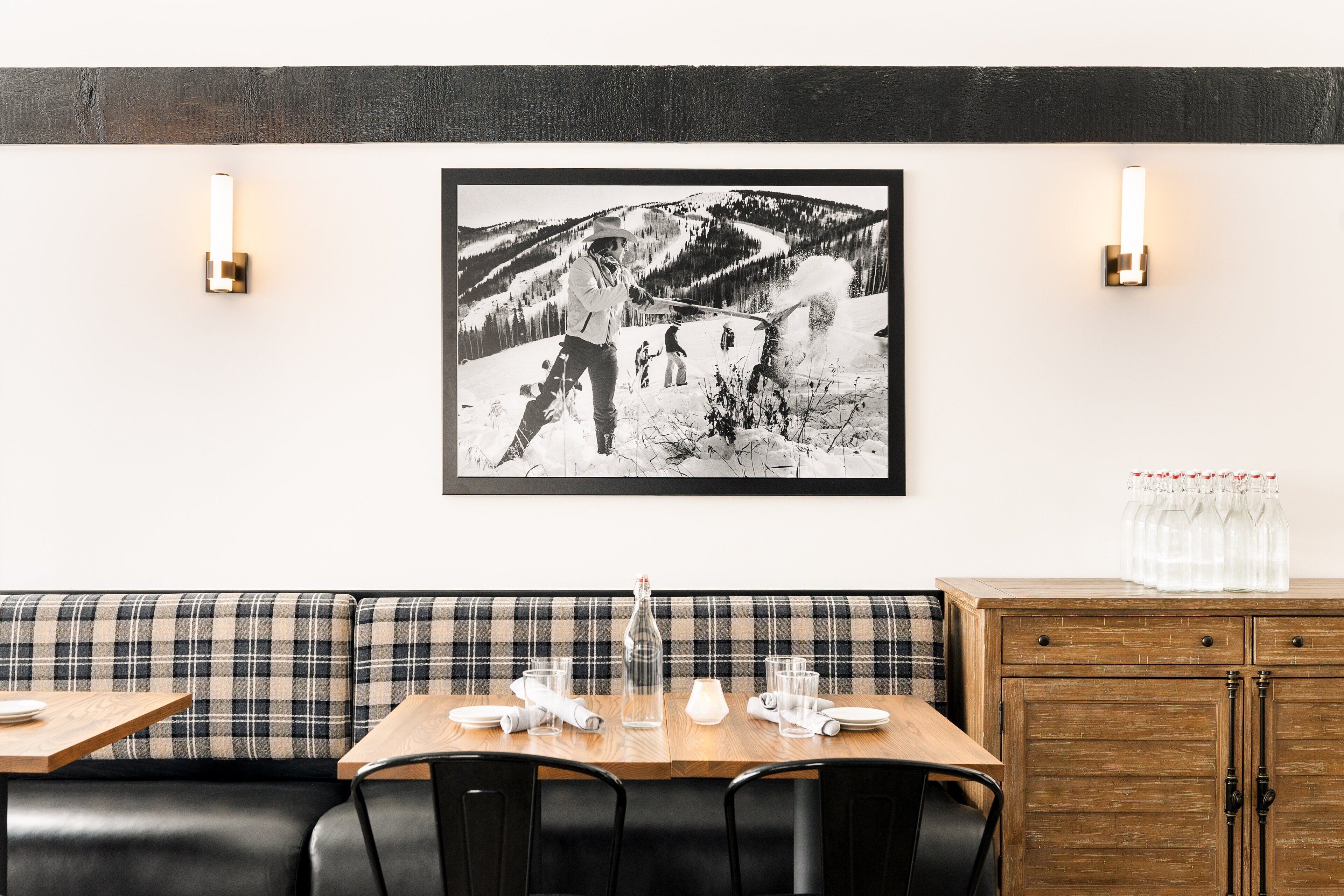 A restaurant with a plaid bench seating area, a wooden table with a bottle and glasses, and a framed black and white photo on the wall.