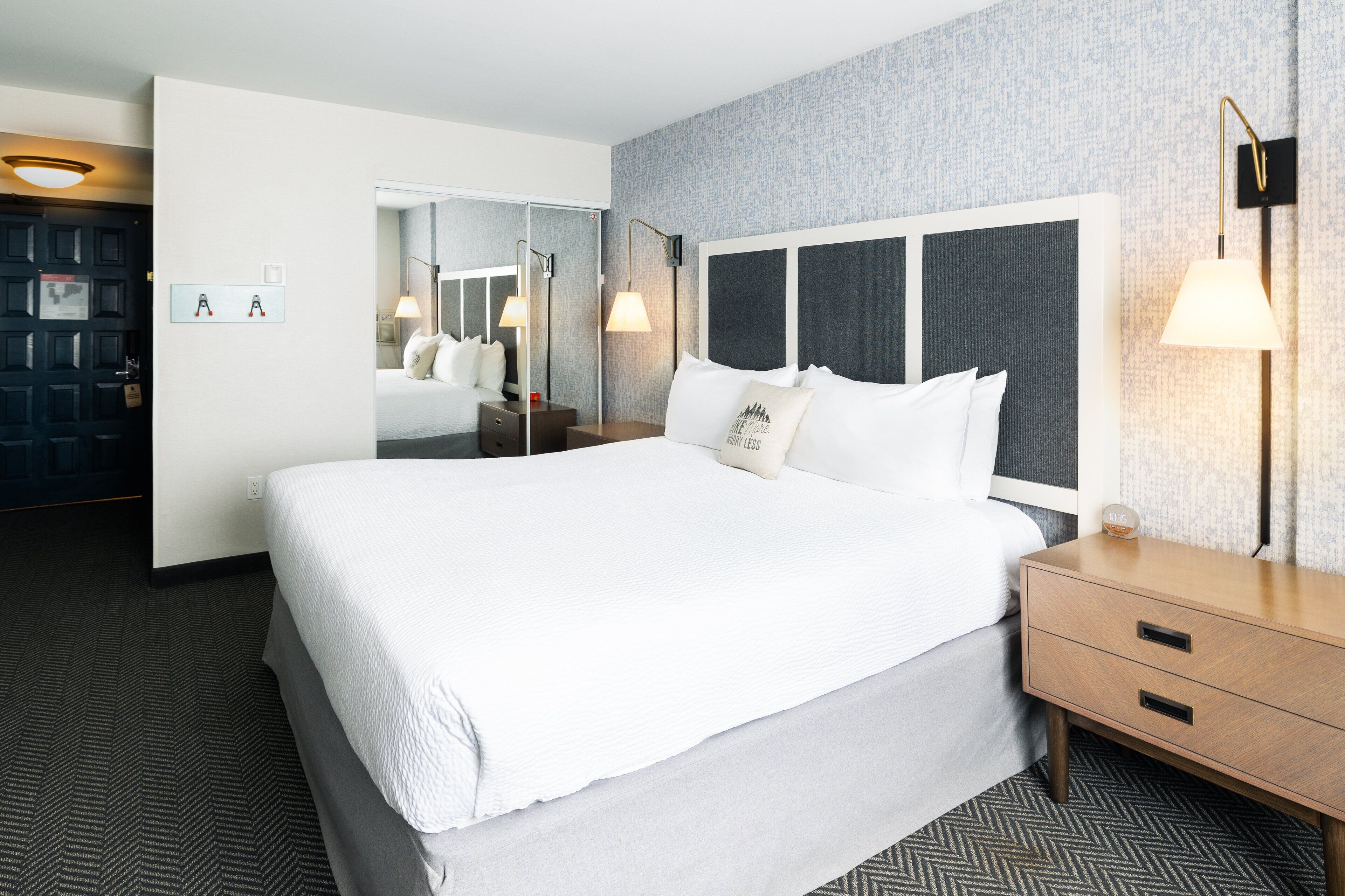 A hotel room with a large bed, a bedside table, a nightstand, a mirror, and a wall-mounted lamp.