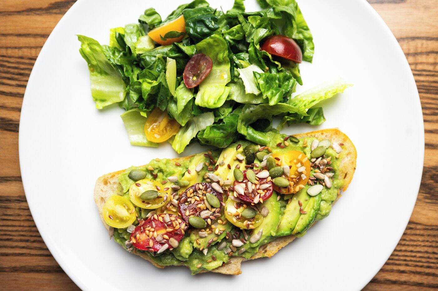A plate with a sandwich topped with avocado and seeds, and a side salad with lettuce and cherry tomatoes.