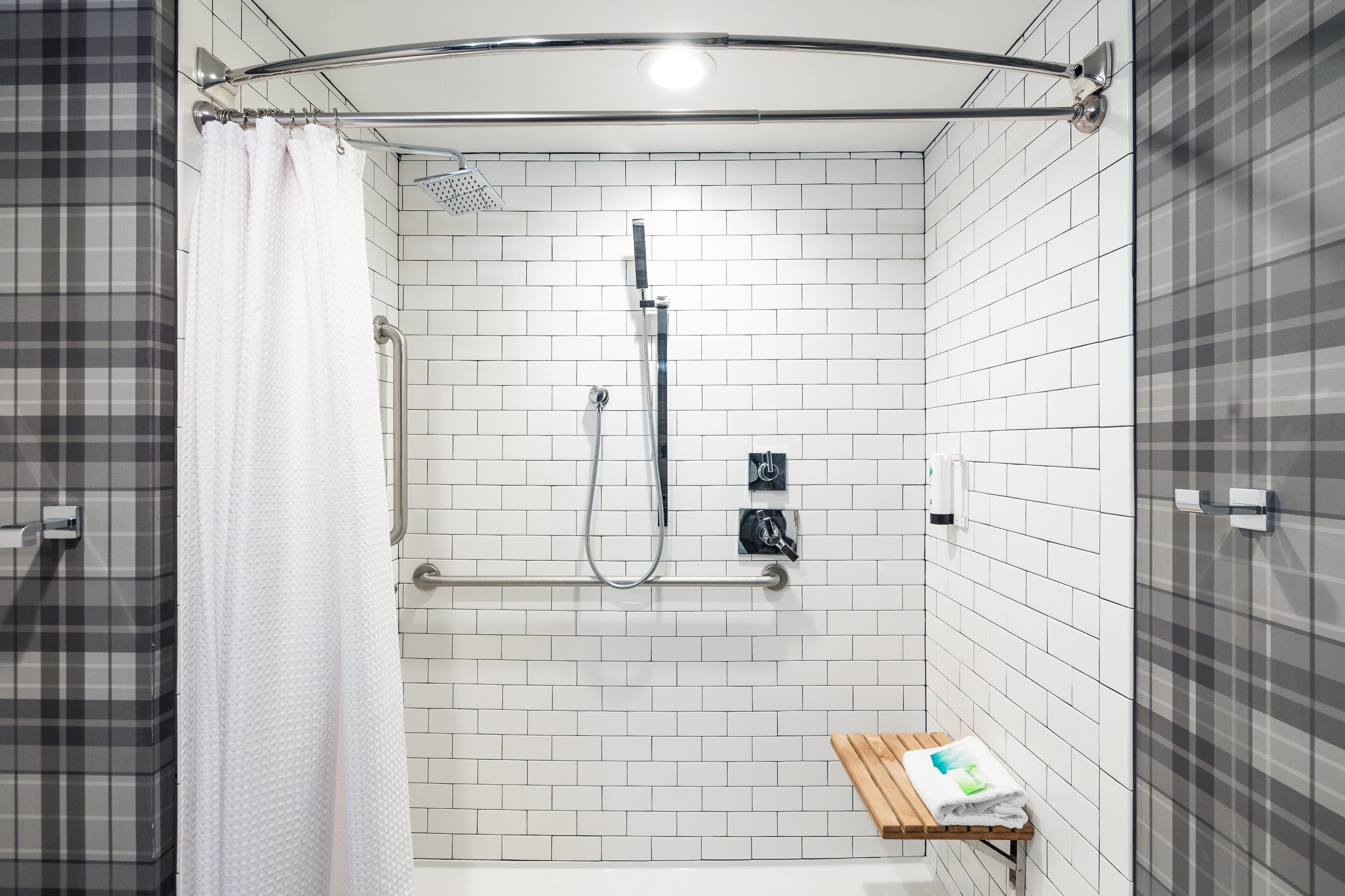 Área de duche com paredes revestidas a azulejos brancos, um chuveiro, um porta-toalhas e um banco com uma toalha.
