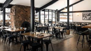 Un restaurant spacieux avec de hauts plafonds, des poutres apparentes et de grandes fenêtres. Les tables sont disposées avec des chaises, et un mur en pierre ajoute une touche rustique.