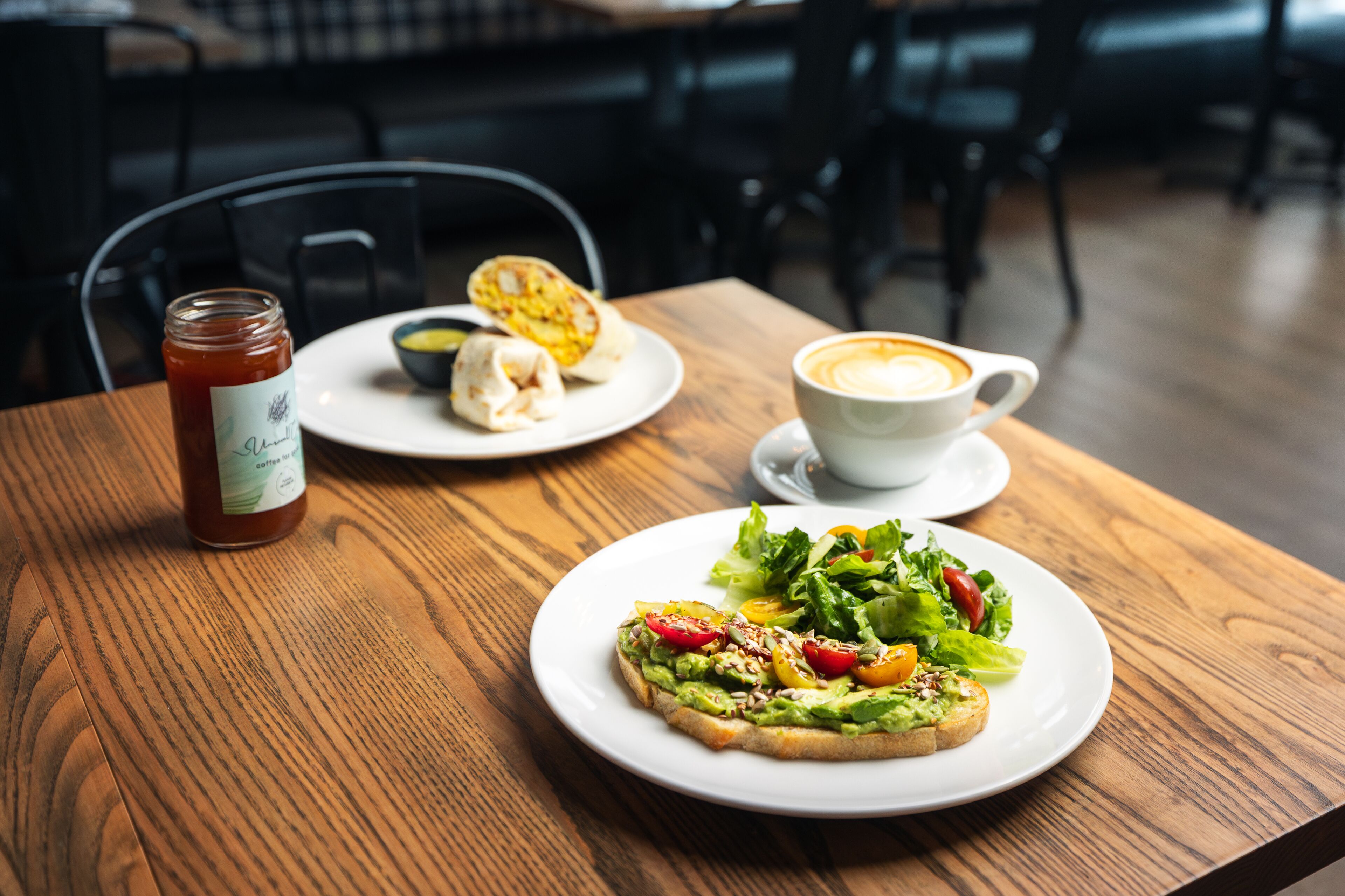 Een houten tafel met avocado-toast, een kop koffie en een pot jam.