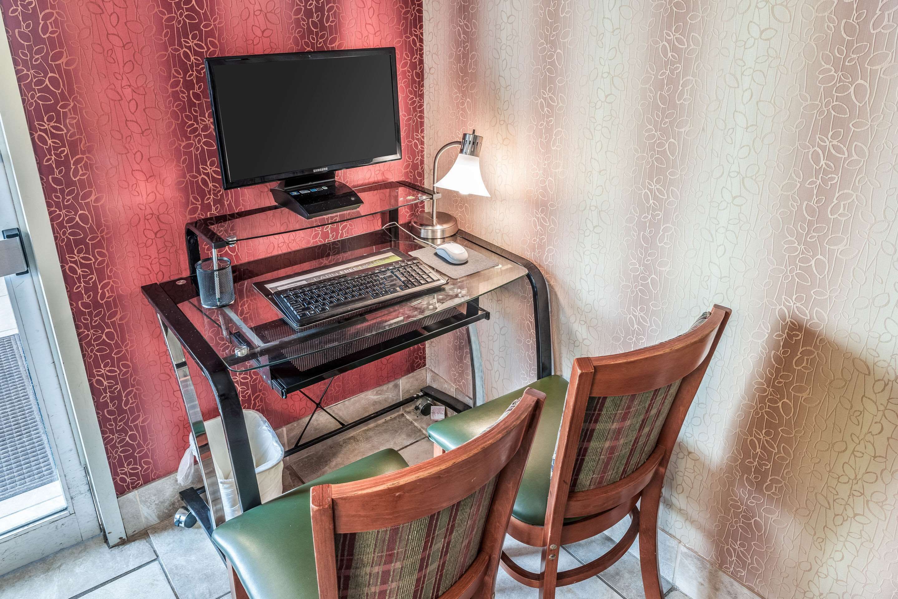 Un bureau en verre avec un ordinateur, une lampe et deux chaises, dans une pièce dont les murs sont recouverts d’un papier peint à motifs.