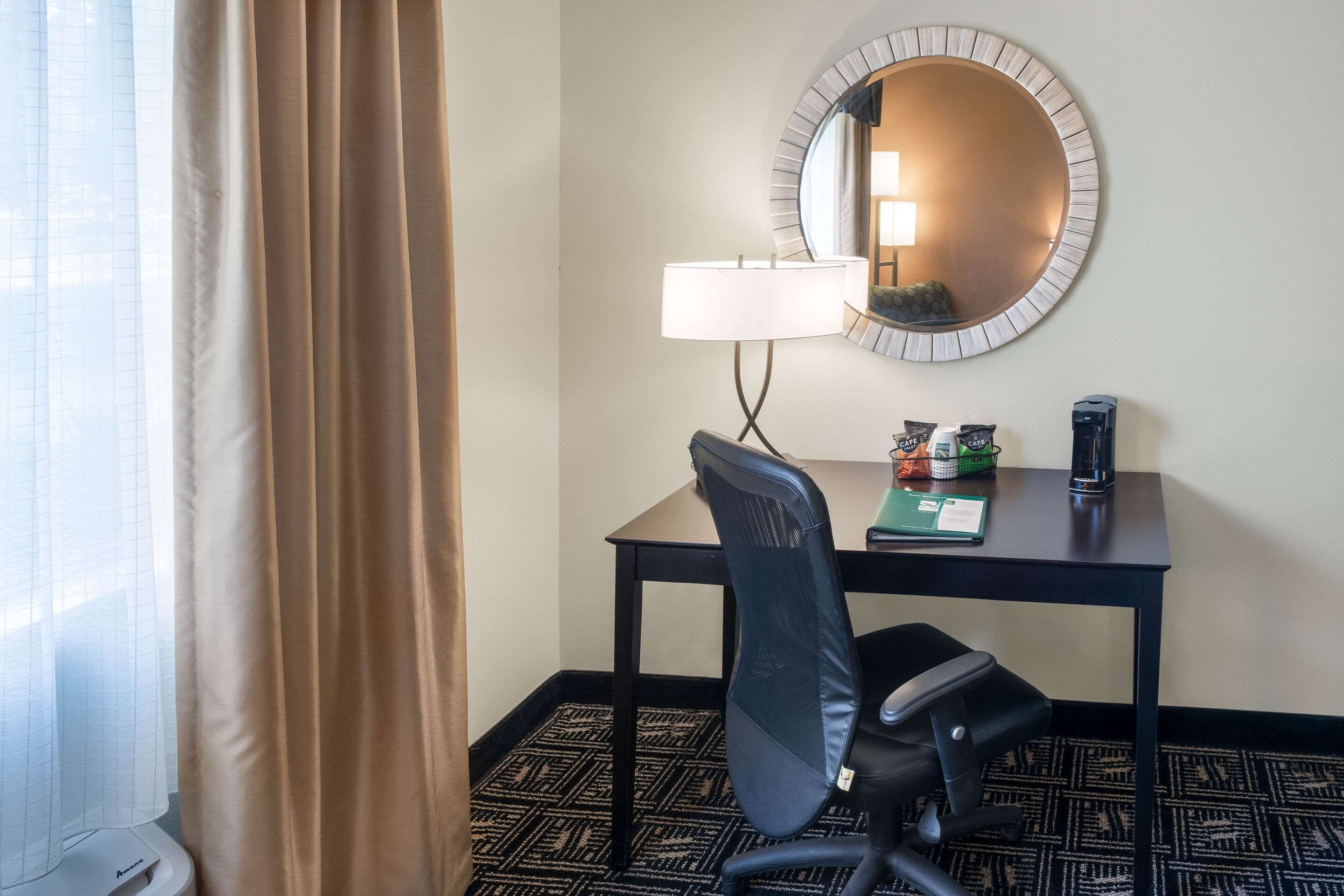 Habitación de hotel con escritorio, silla, lámpara y un espejo redondo en la pared.