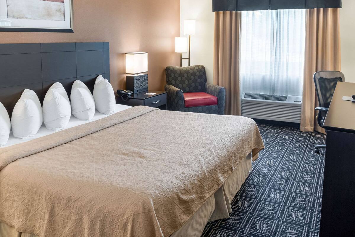 A hotel room with a large bed, a chair with a red cushion, a desk with a computer, and a window with curtains.