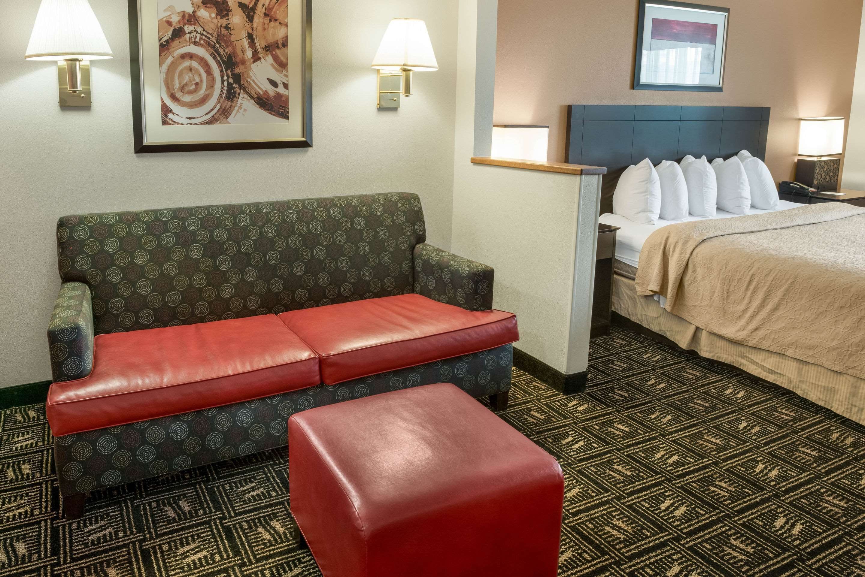 Quarto de hotel com um sofá verde estampado, um banquinho vermelho, uma cama com almofadas brancas e um tapete estampado.