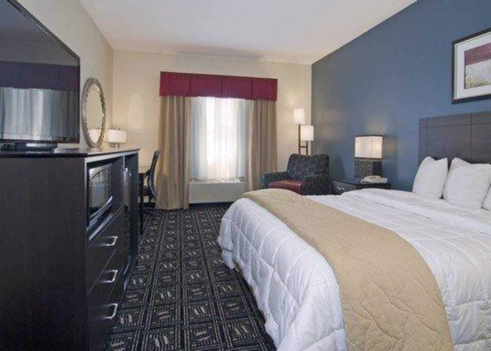 Una habitación de hotel con cama, escritorio, silla, televisor y ventana con cortinas.