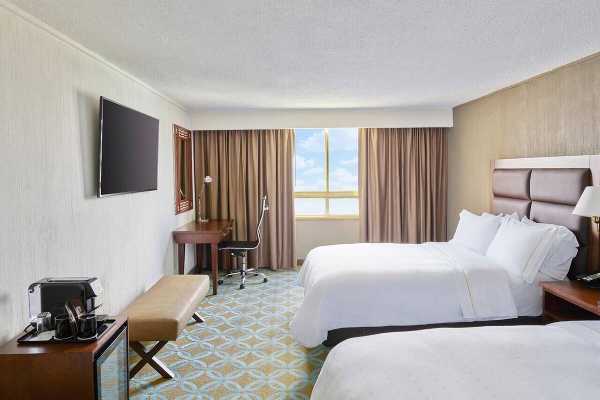 A hotel room with two beds, a desk with a chair, a TV mounted on the wall, and a window with curtains.