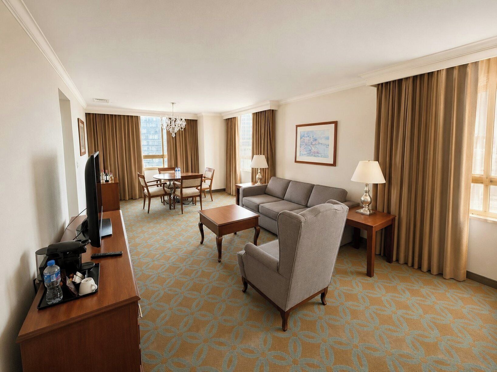 A hotel room with a sofa, armchair, dining table, and a TV on a desk.