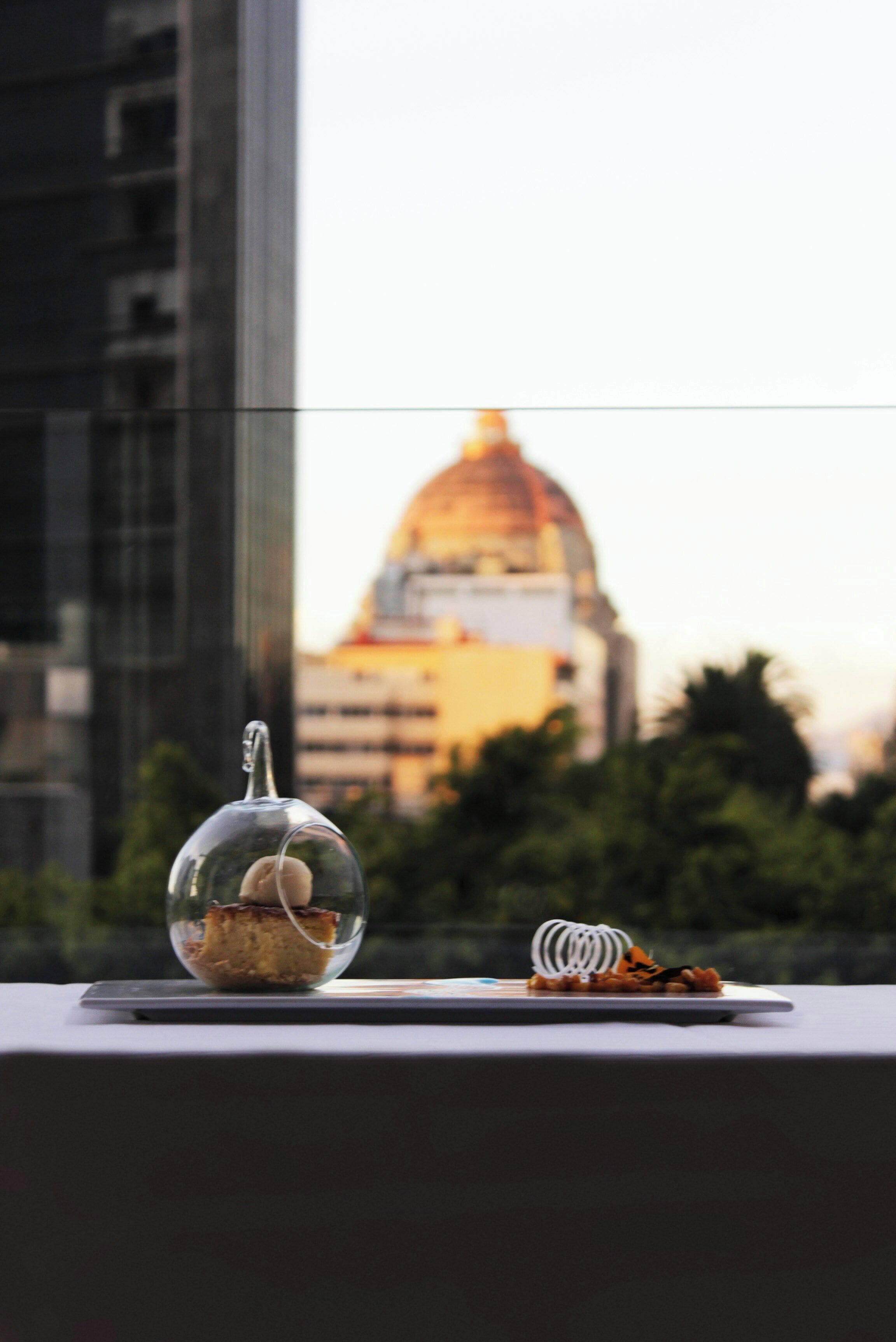 Una cúpula de cristal que contiene un pastel y un muelle enrollado sobre una mesa, con un paisaje urbano al fondo.