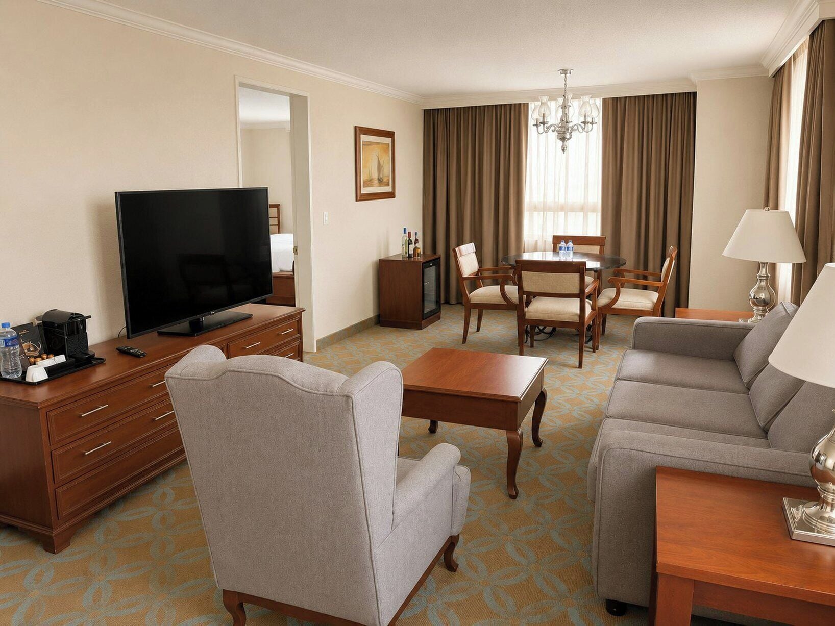A hotel room with a flat-screen TV, a sofa, a chair, a small table, a dining table with chairs, a chandelier, and a painting on the wall.