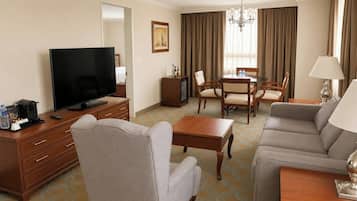 A hotel room with a flat-screen TV, a sofa, a chair, a small table, a dining table with chairs, a chandelier, and a painting on the wall.