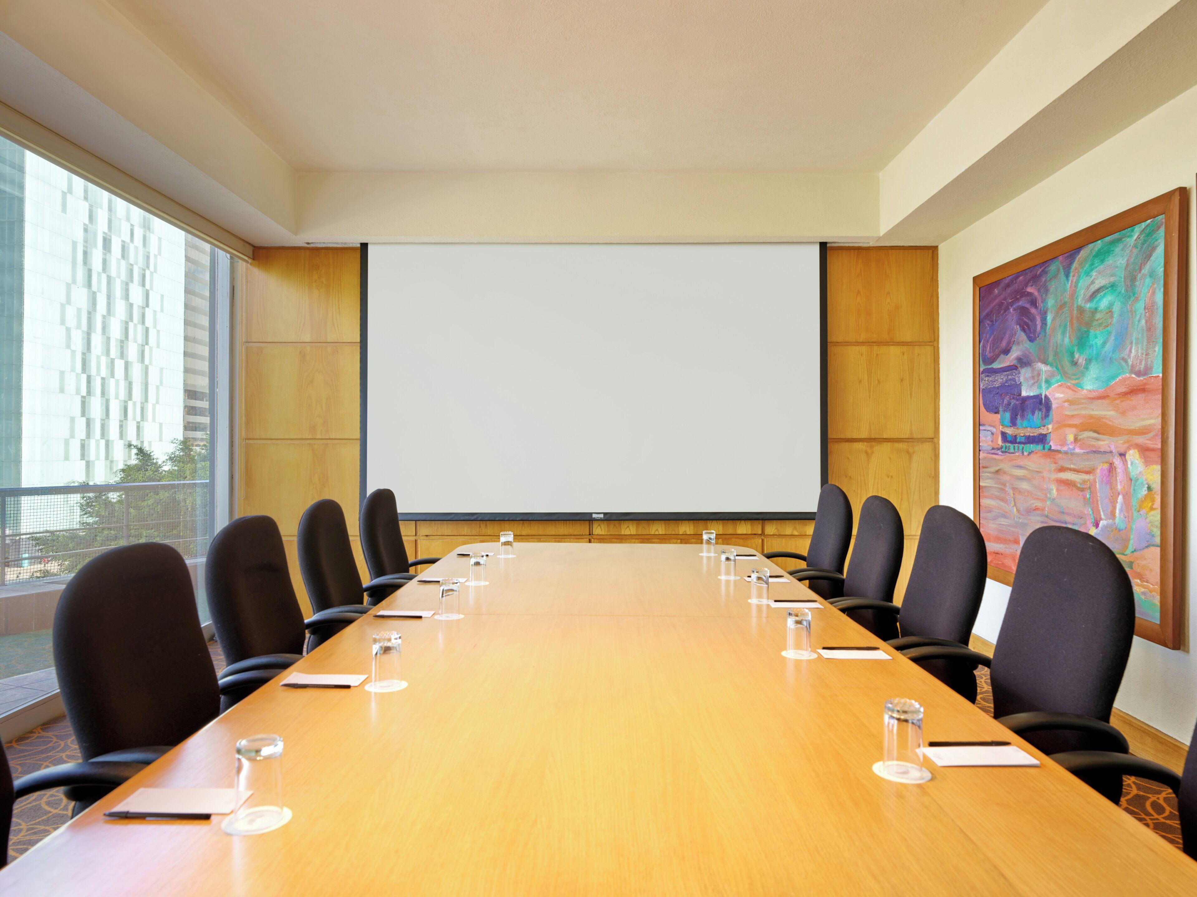 A conference room with a long table, chairs, a projector screen, and a painting on the wall.