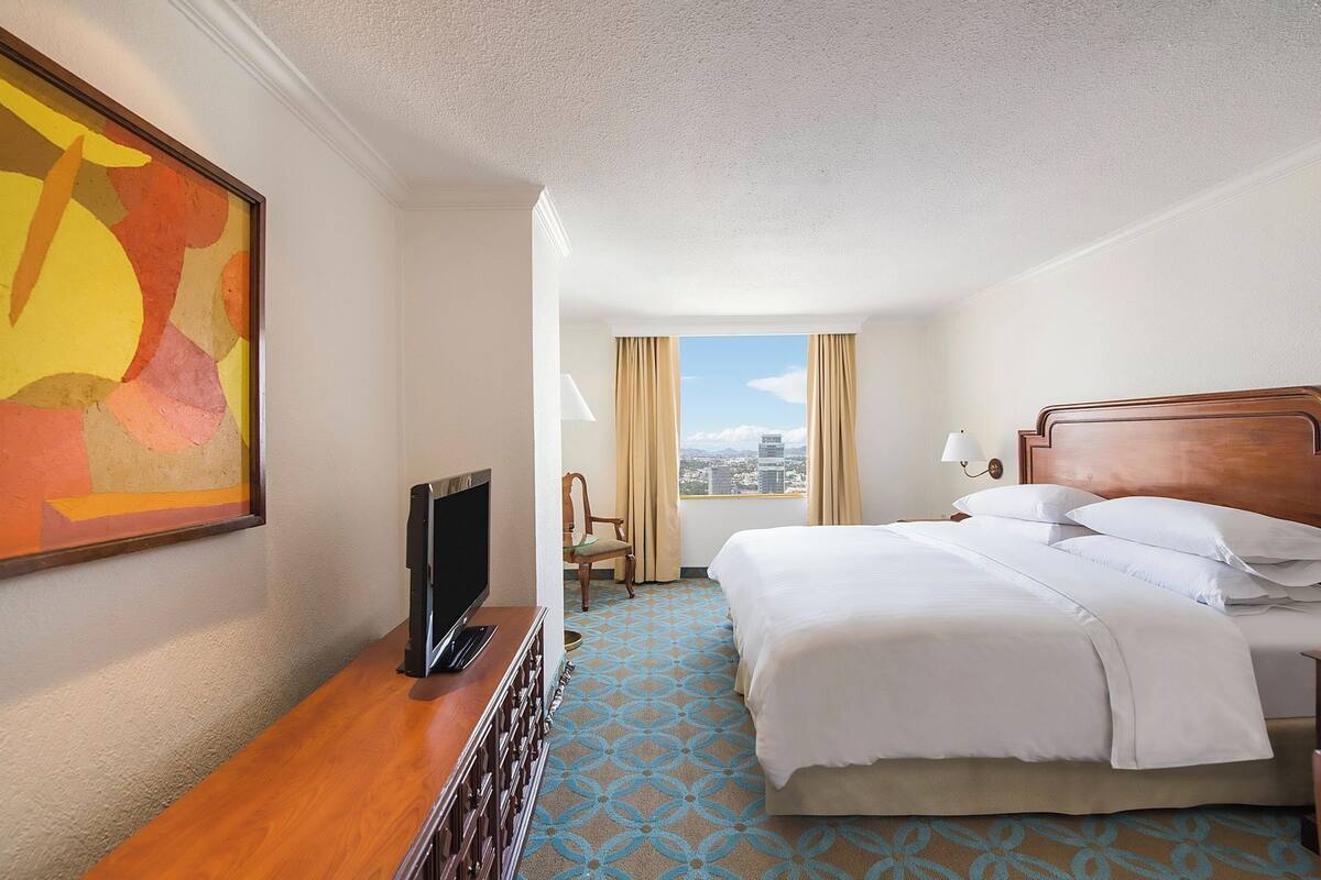 A hotel room with a bed, a TV, a chair, a window with a city view, and a painting on the wall.