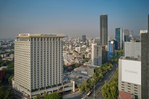 Un hôtel de grande hauteur, reconnaissable à son grand panneau « FESTA AMERICA », s’élève au milieu d’un paysage urbain dense, composé de différents bâtiments, sous un ciel dégagé.