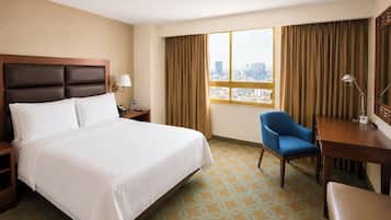 A hotel room with a bed, a desk, a chair, a lamp, and a window with a city view.