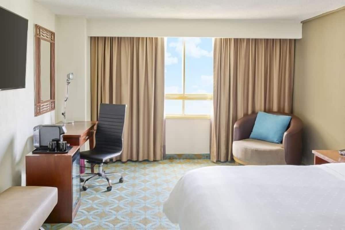 A hotel room with a bed, a desk with a chair, a window with curtains, and a view of the sky.