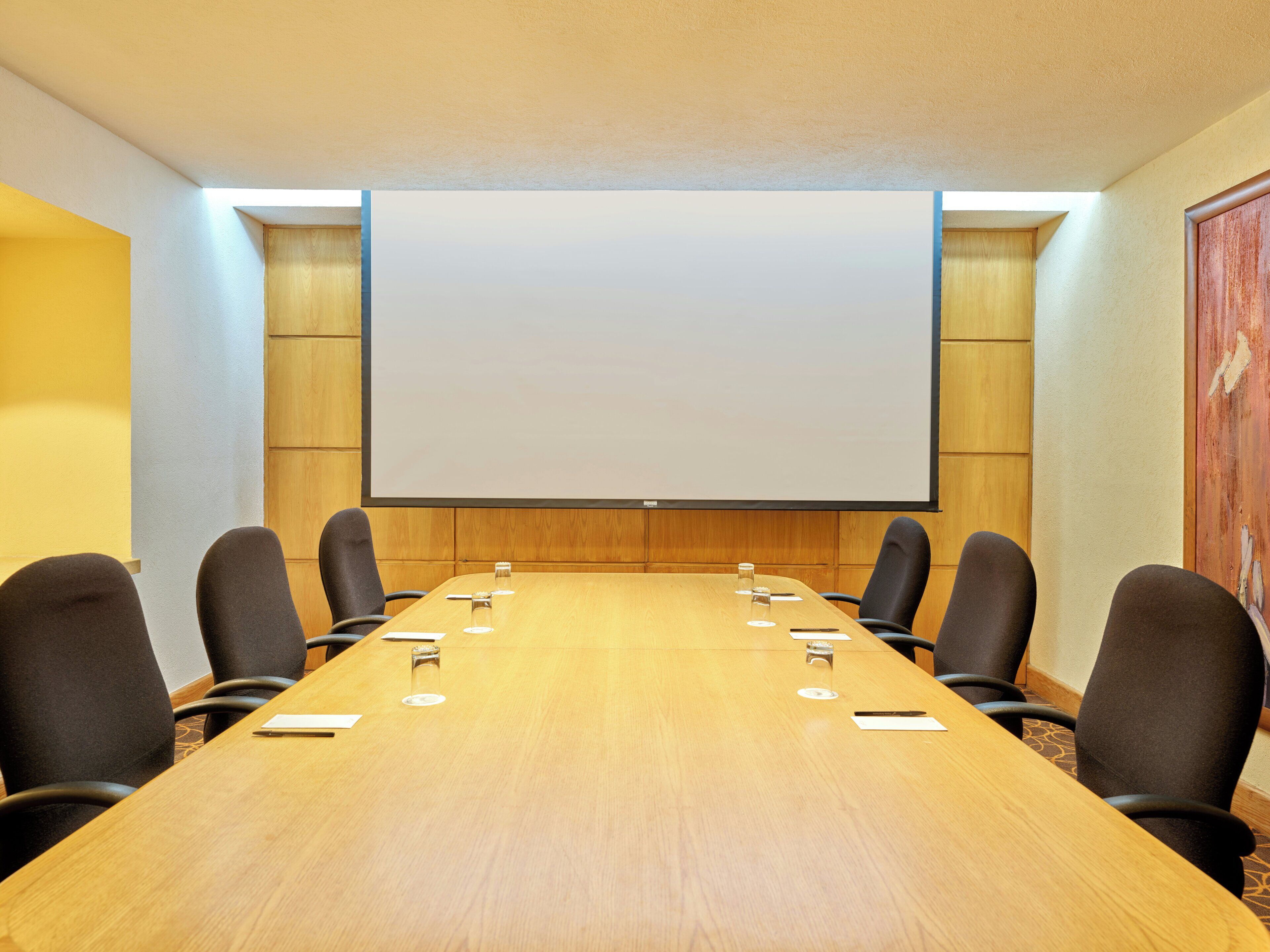 Un salón de conferencias con una mesa larga, seis sillas, una pantalla de proyector y un revestimiento de pared de madera.