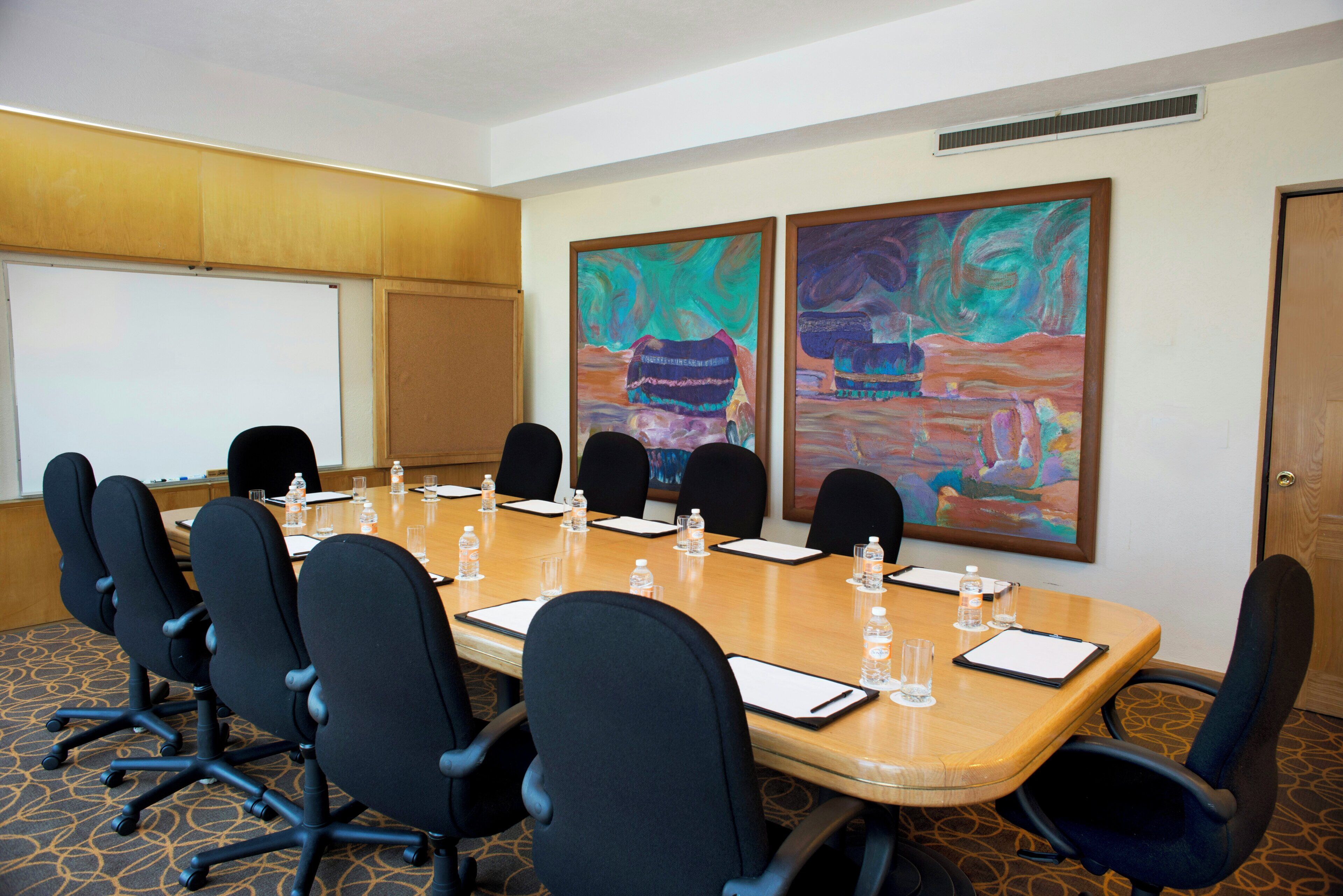 A conference room with a long table, black chairs, water bottles, and two large abstract paintings on the wall.