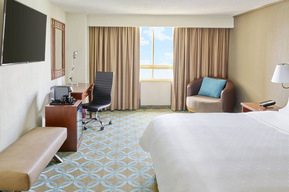 A hotel room with a bed, a desk with a chair, a sofa, a TV, and a window with curtains.