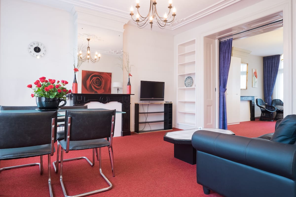 Ein modernes Wohnzimmer mit einer schwarzen Ledersofa, einem Esstisch mit Stühlen, einem Fernseher und einem roten Teppich.