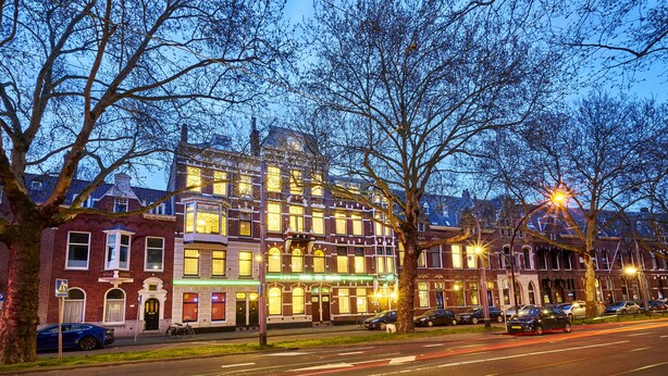 A row of Victorian-style buildings with illuminated windows and a street lined with parked cars and trees.