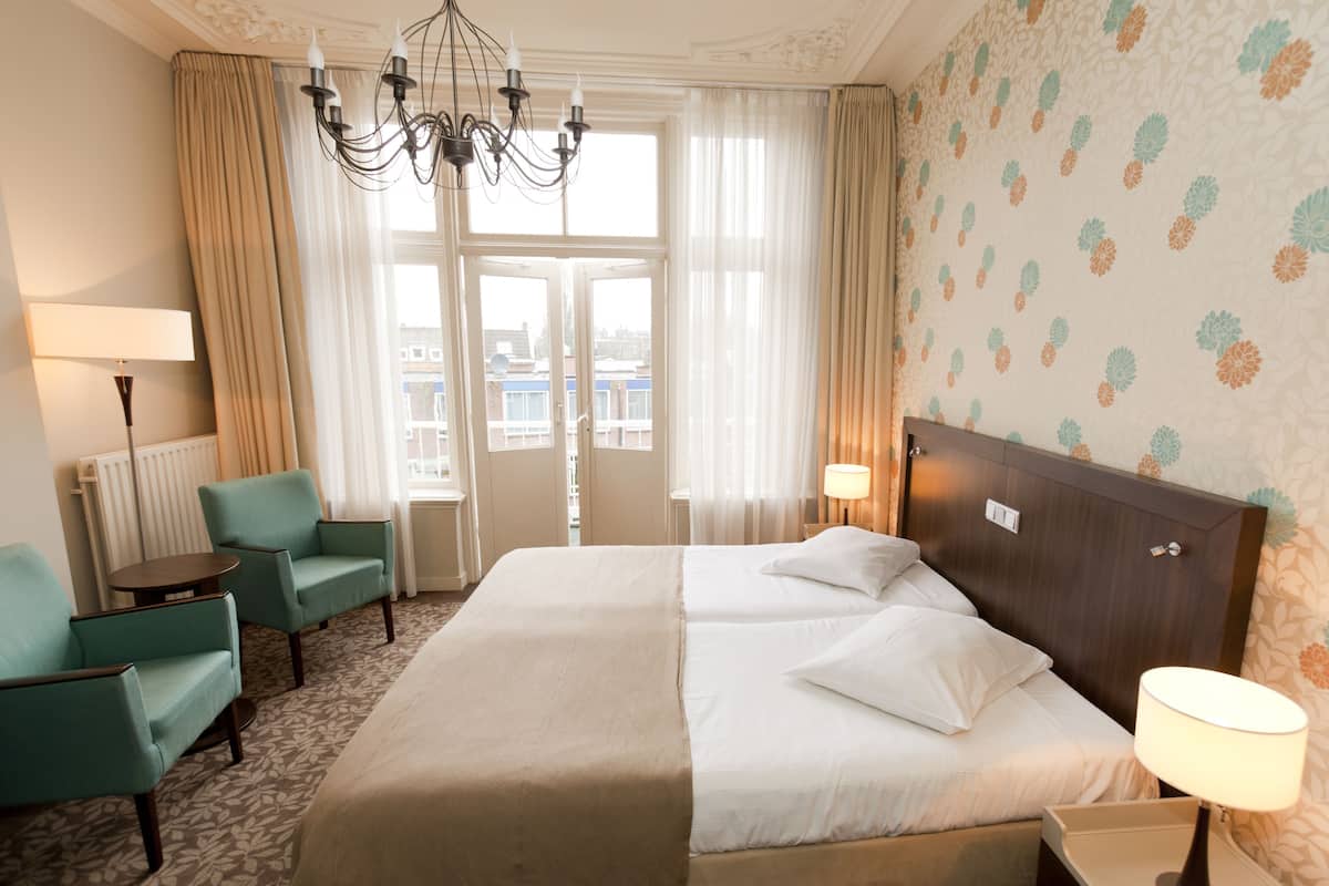 Ein Hotelzimmer mit einem großen Bett, zwei Sesseln, einem kleinen Tisch, einer Lampe und Blick auf die Stadt durch große Fenster.
