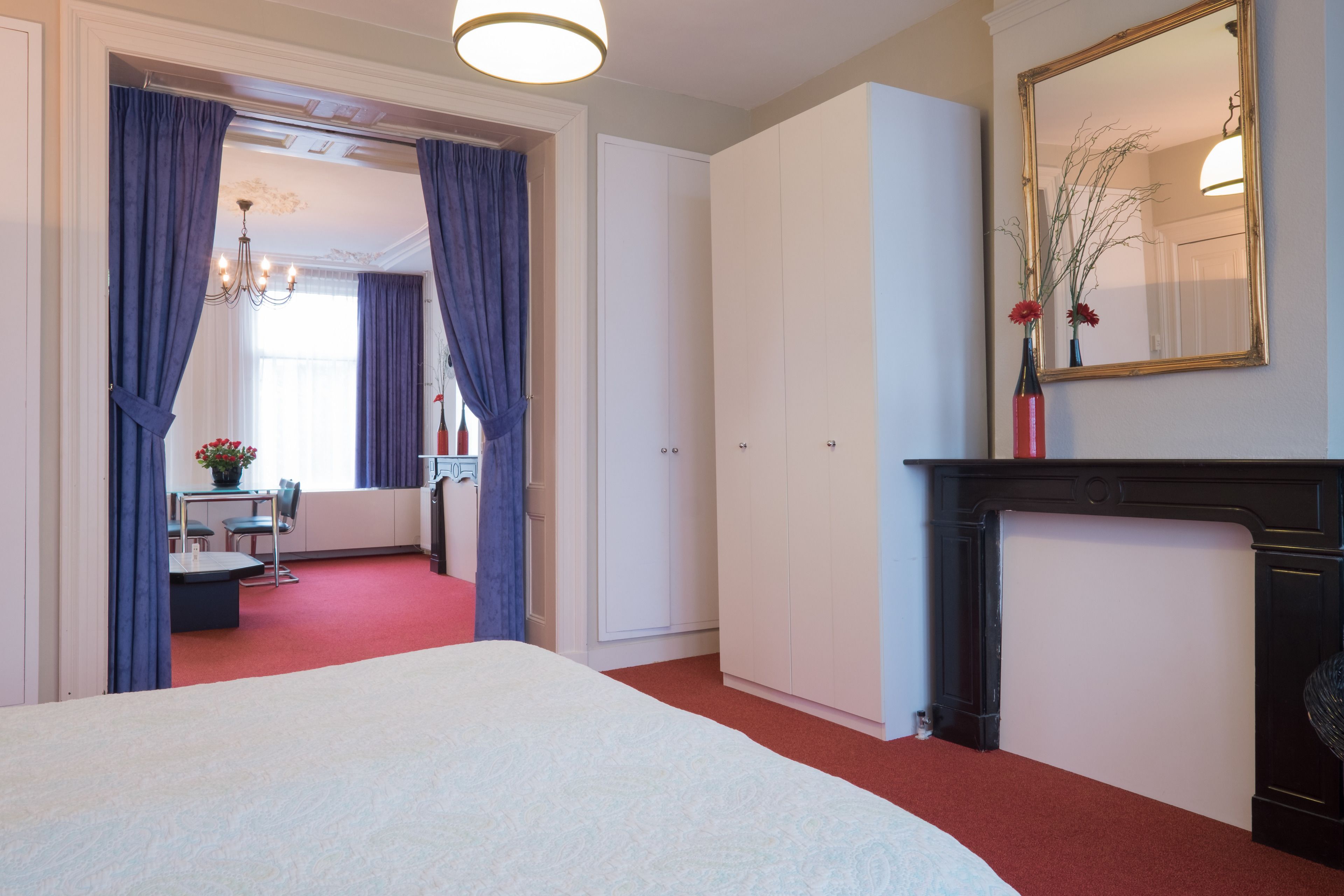 A hotel room with a large bed, a fireplace, a dining area with a table and chairs, and a mirror on the wall.