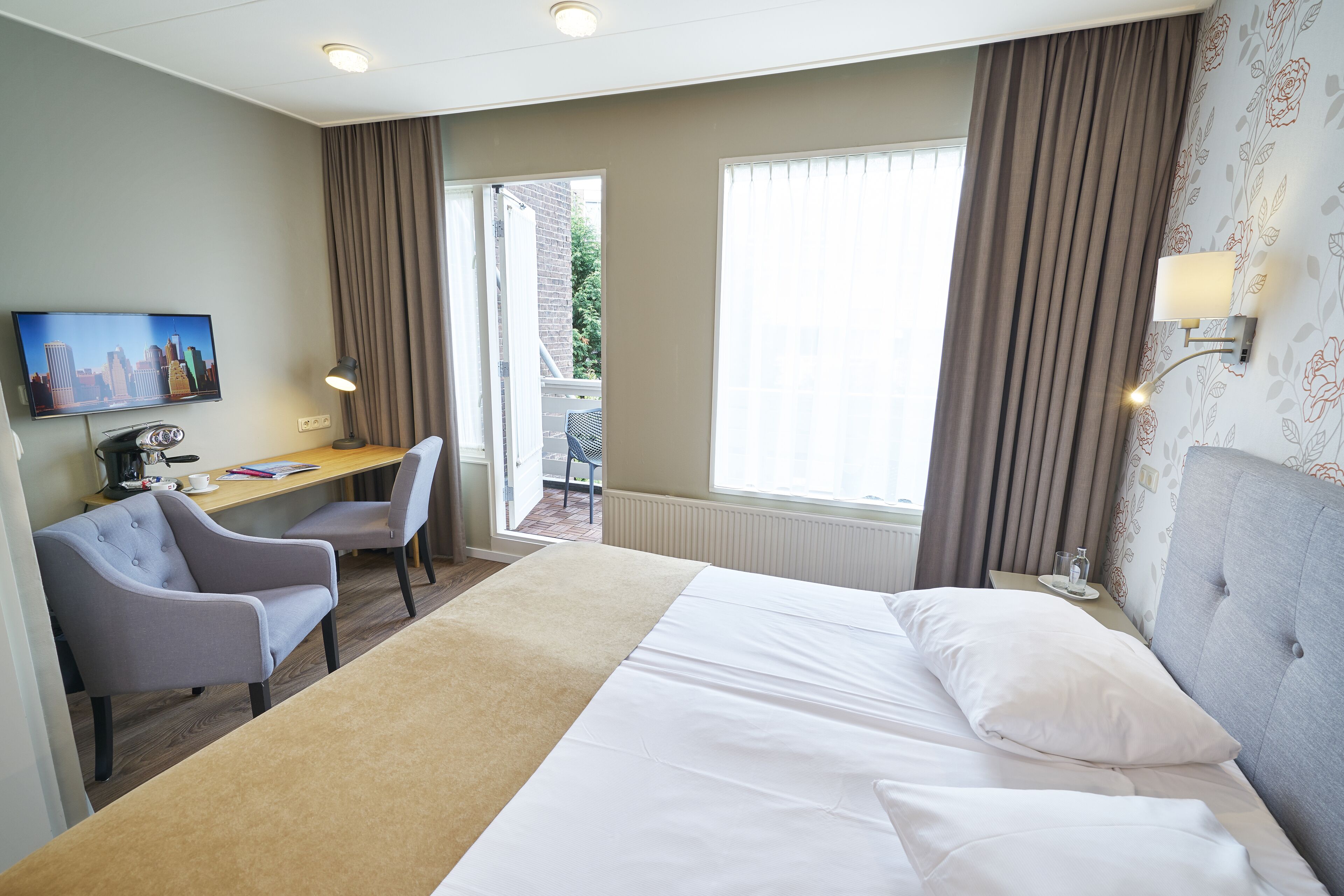 Habitación de hotel con cama, escritorio, silla, televisión y ventana con cortinas.