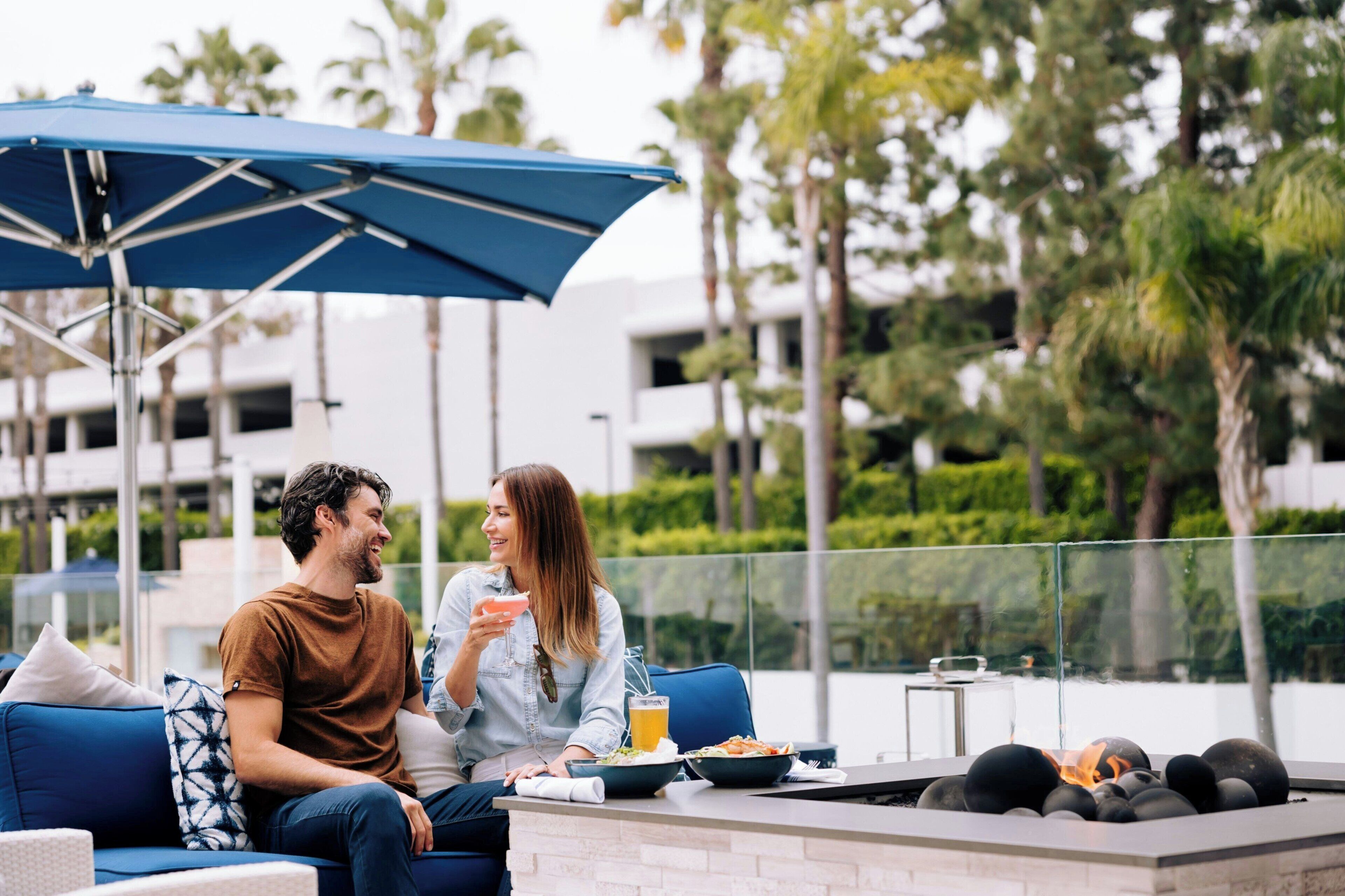 A couple dining at an outdoor patio with a fire feature, blue furniture, and a view of a modern building and palm trees.