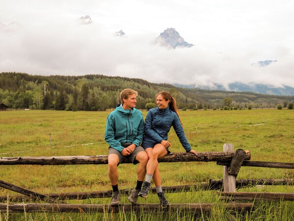 Dos personas sentadas sobre una valla de madera en un campo cubierto de hierba, con montañas al fondo.