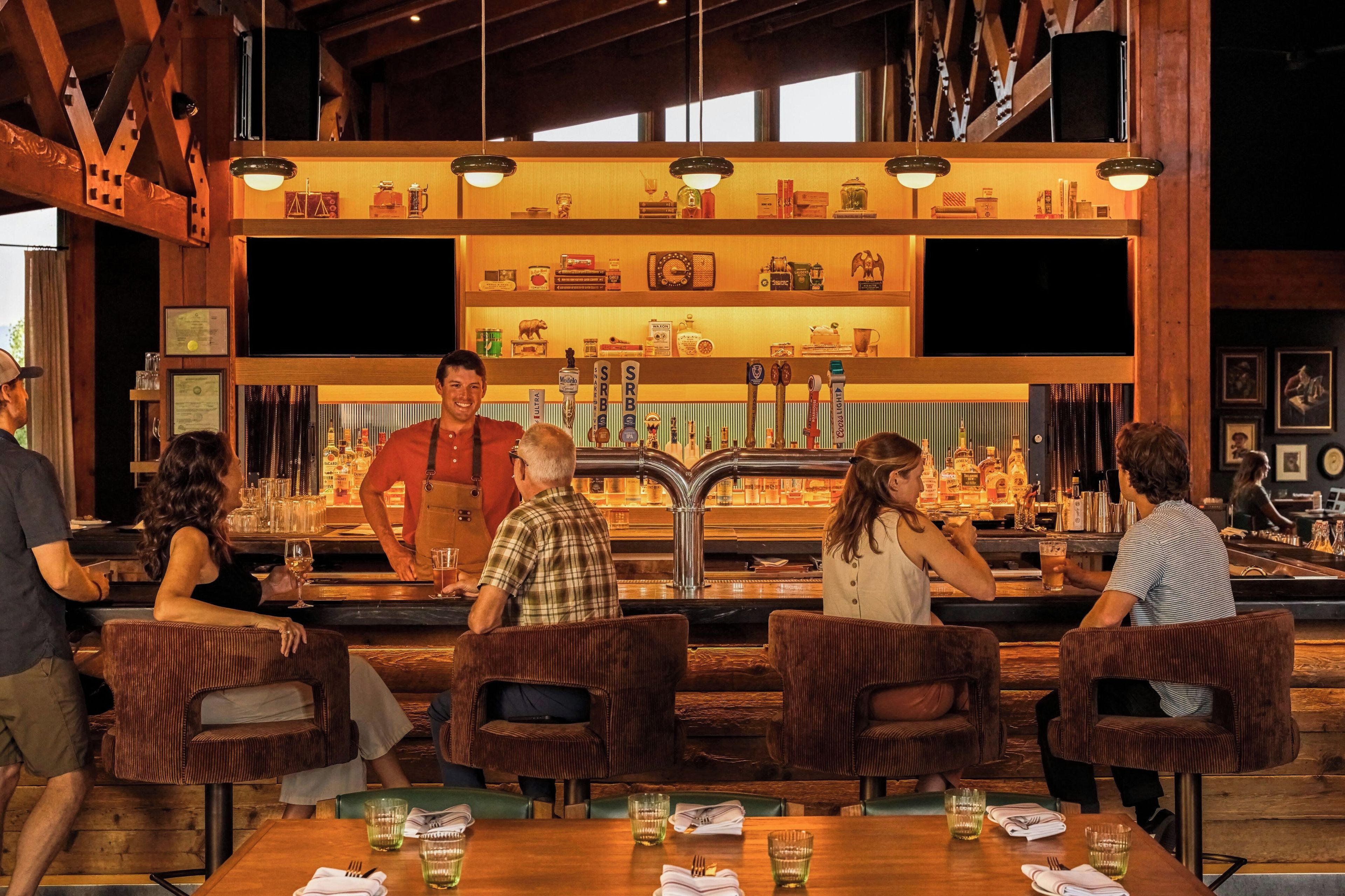 A bar area with wooden furniture, a bar counter with various bottles, and a person serving drinks.