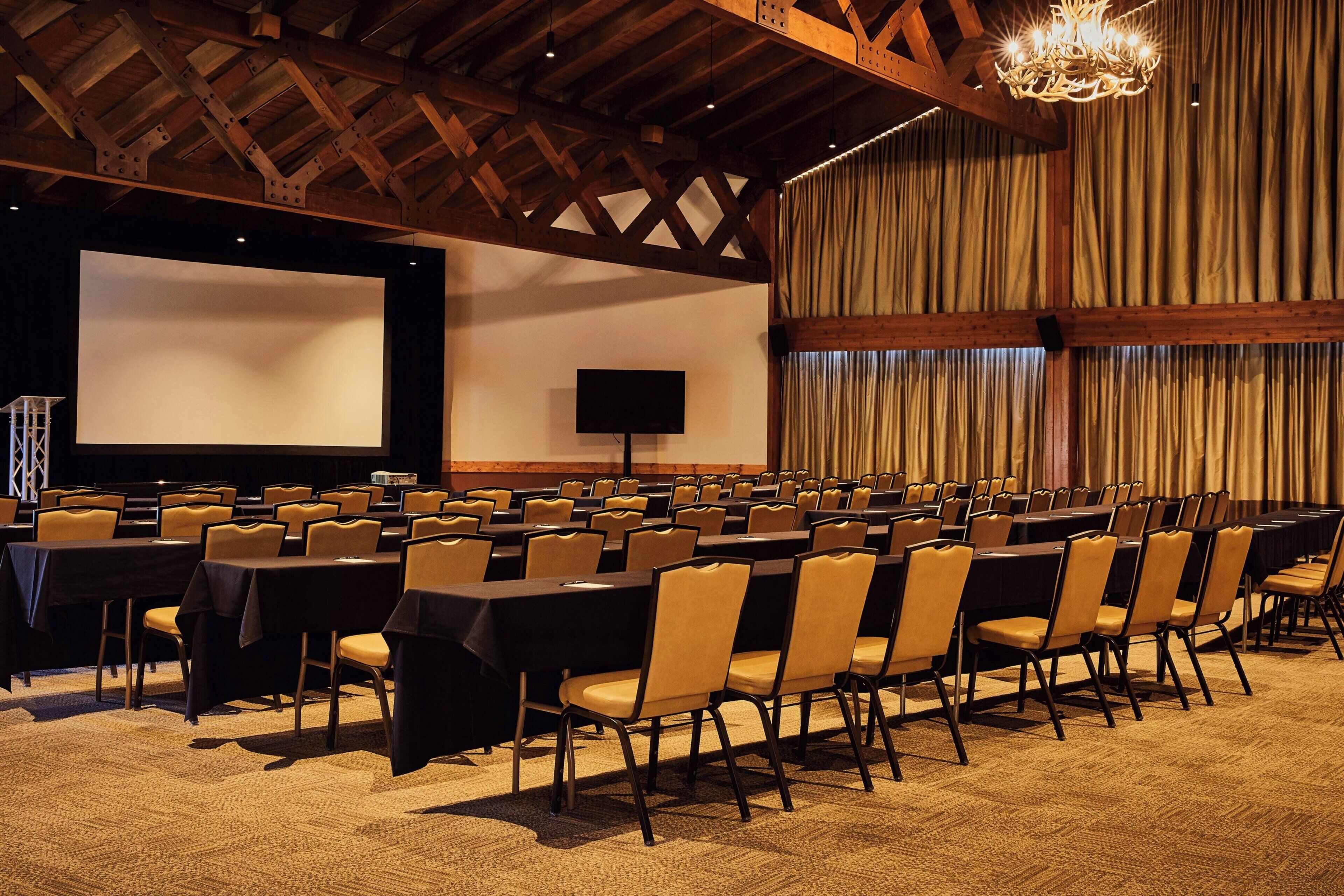A conference room with rows of tables and chairs, a large screen, and a speaker.