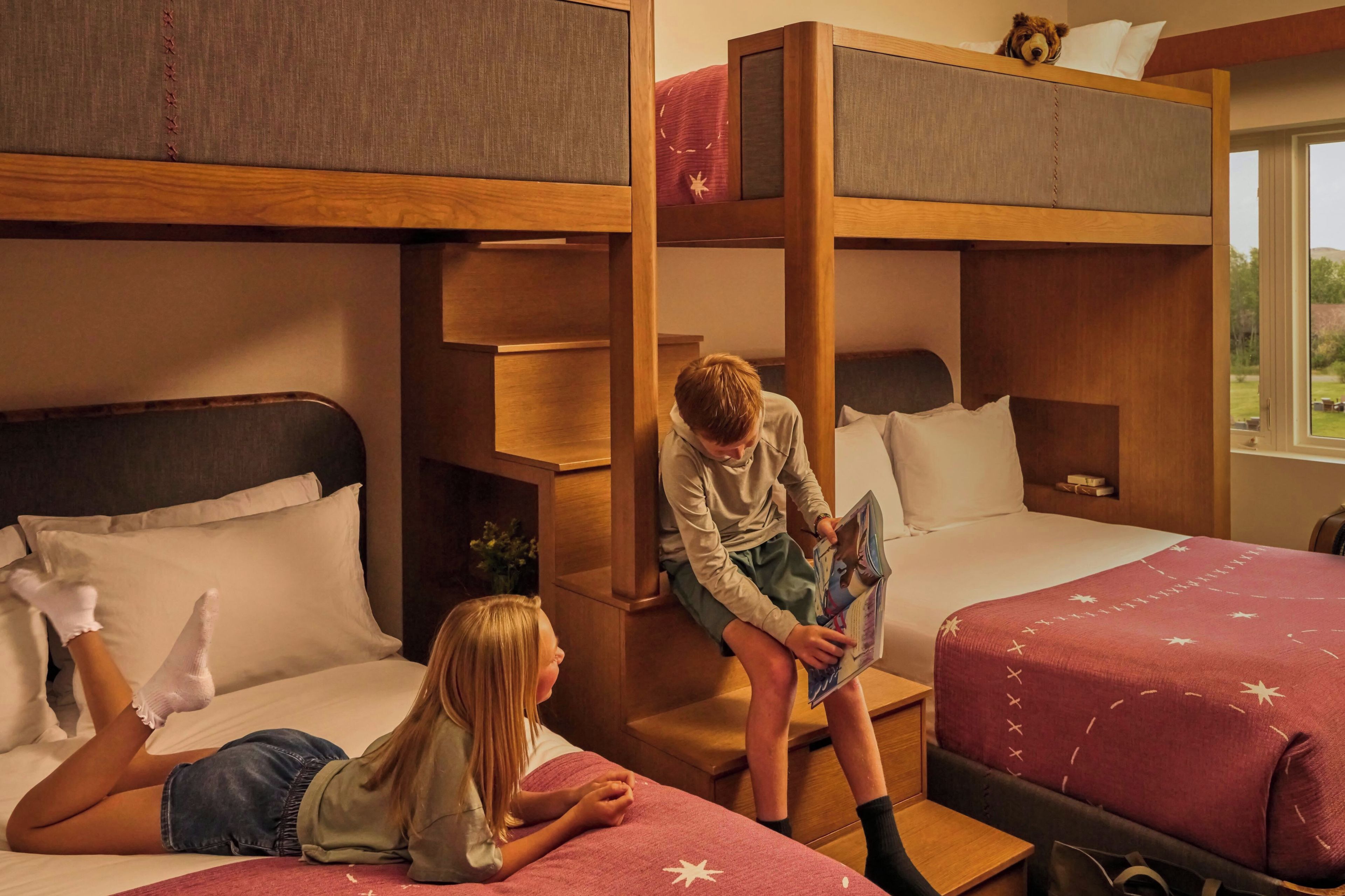A dormitory room with bunk beds, a small table, and a window with a view of the outdoors.