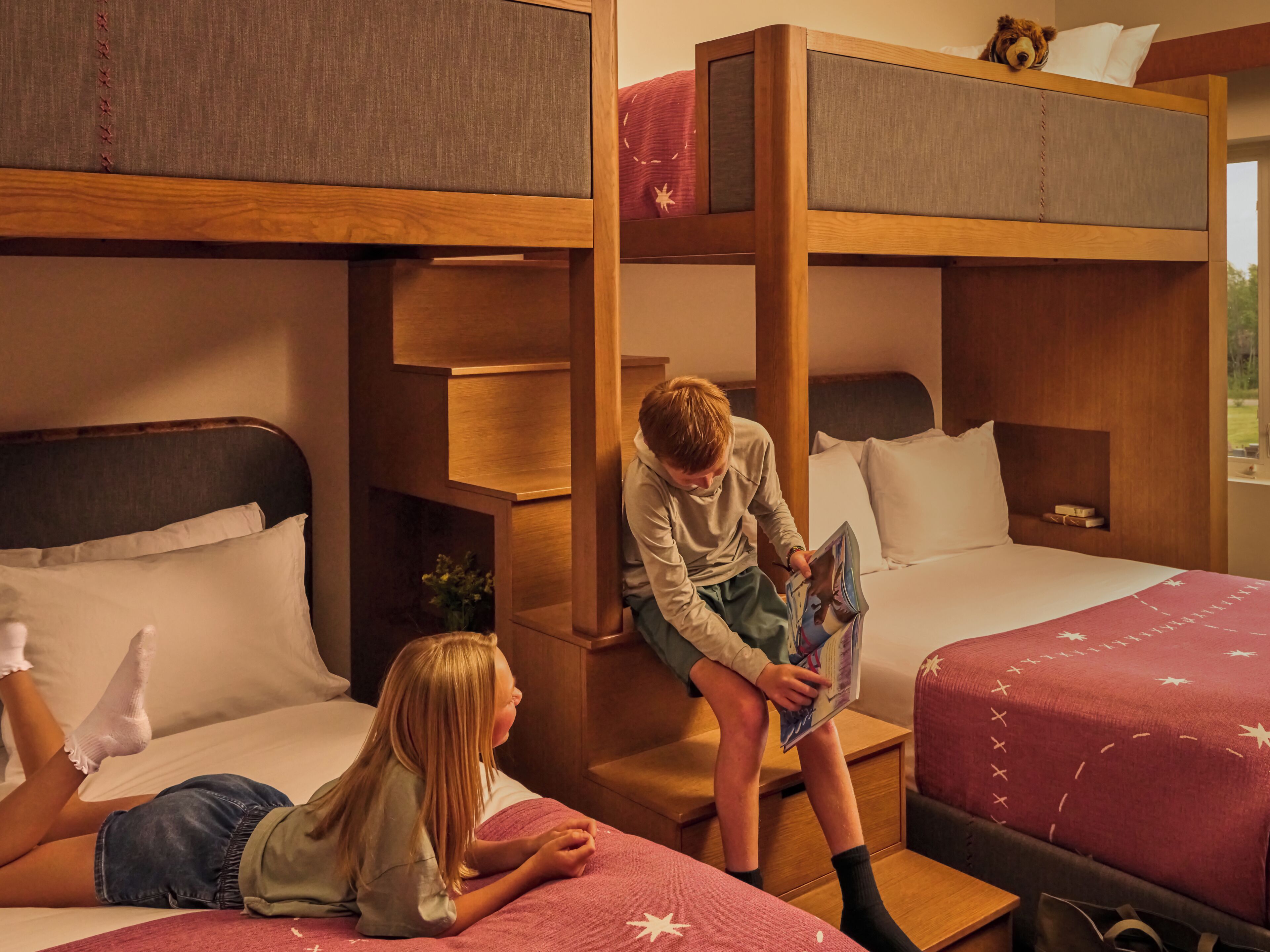 A bunk bed room with a wooden ladder, a lower bunk with a pillow, and a top bunk with a teddy bear.