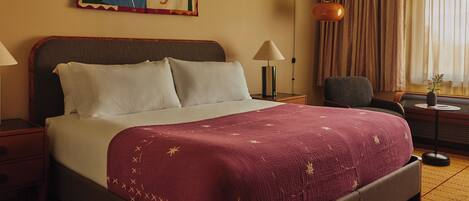Une chambre d’hôtel avec un lit, une table de chevet, une lampe, une chaise et une fenêtre avec des rideaux.