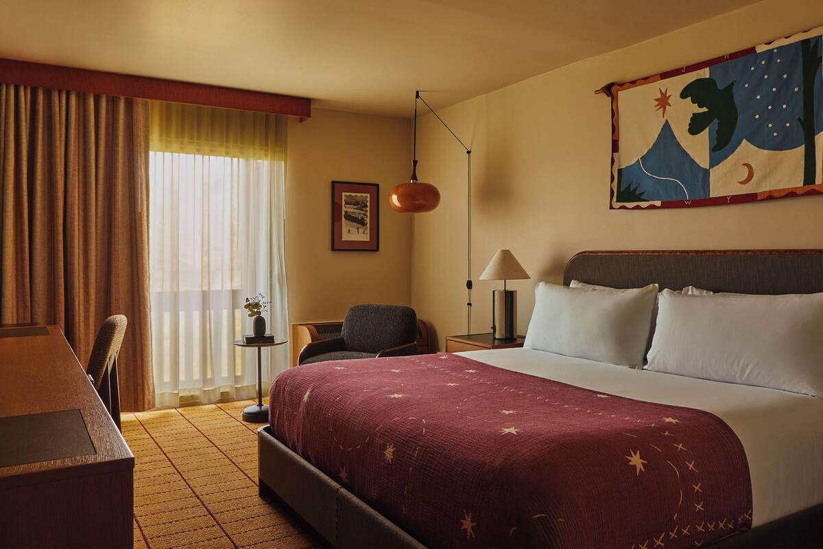 A hotel room with a bed, a desk, a chair, a lamp, a painting, and a window with curtains.