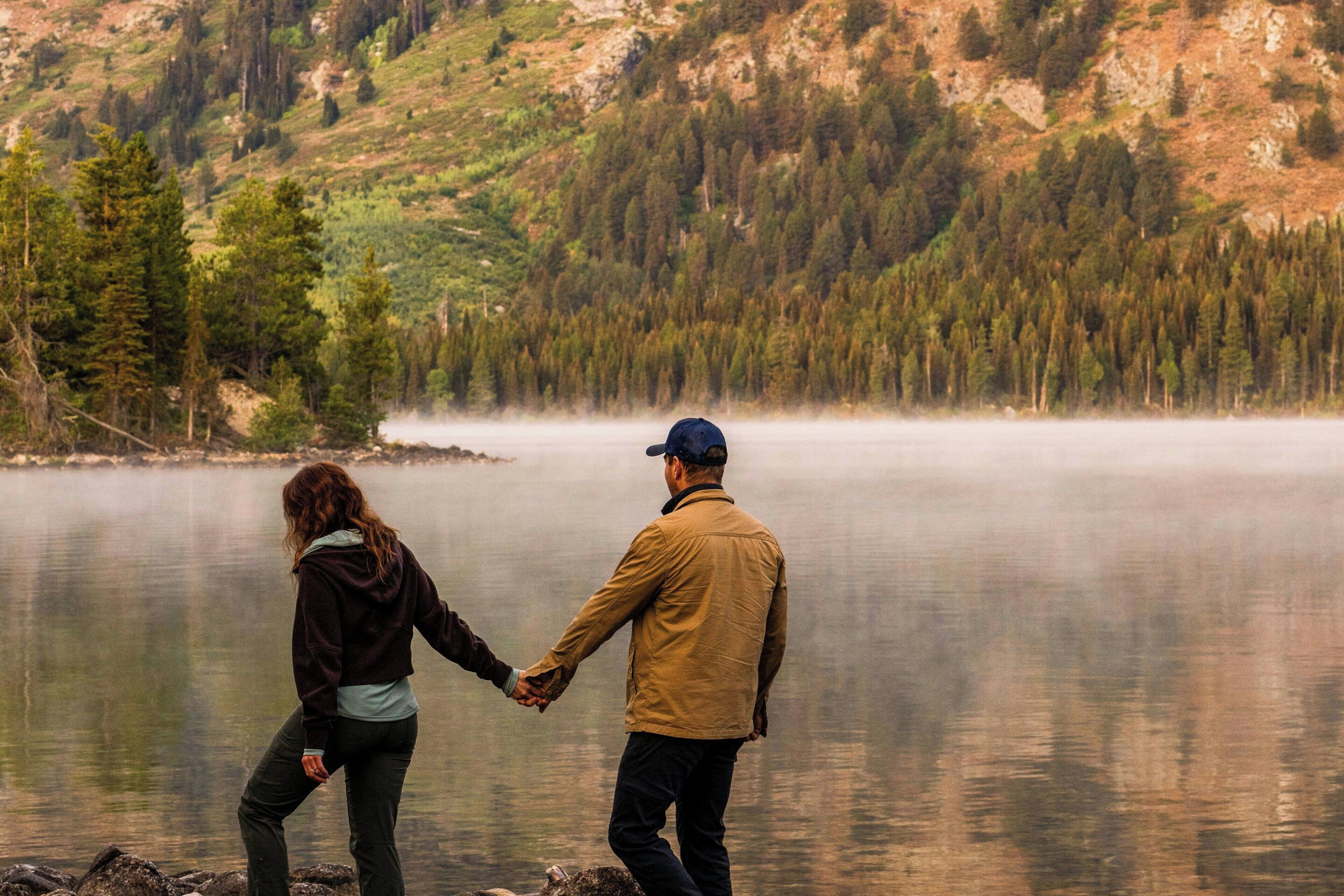 A couple holding hands by a calm lake with a forested mountain backdrop.