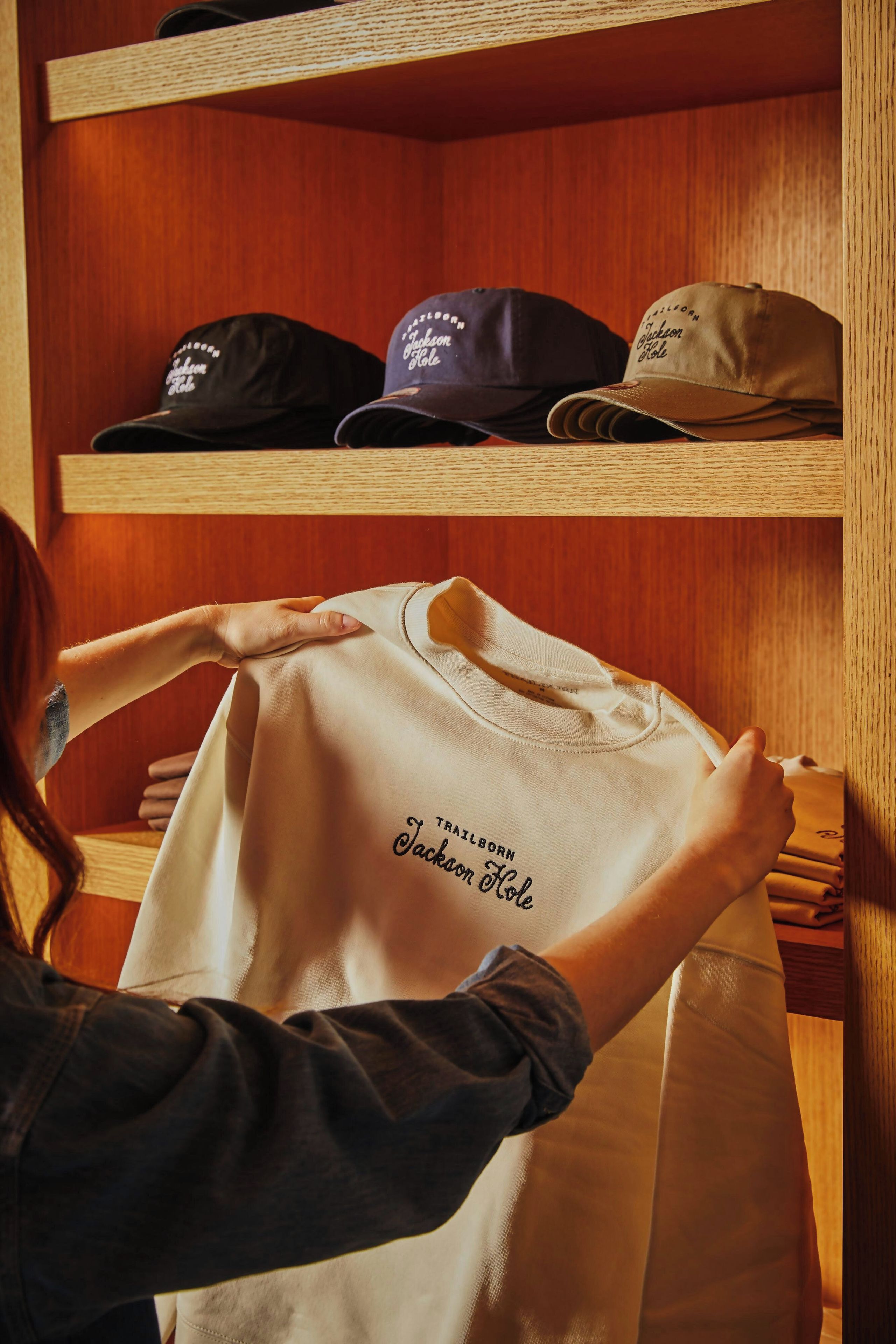 Une personne porte un t-shirt blanc sur lequel on peut lire « TRAILBORN JACKSON HOLE », et au-dessus, des étagères en bois sont garnies de chapeaux.