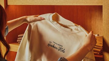 Une personne porte un t-shirt blanc sur lequel on peut lire « TRAILBORN JACKSON HOLE », et au-dessus, des étagères en bois sont garnies de chapeaux.