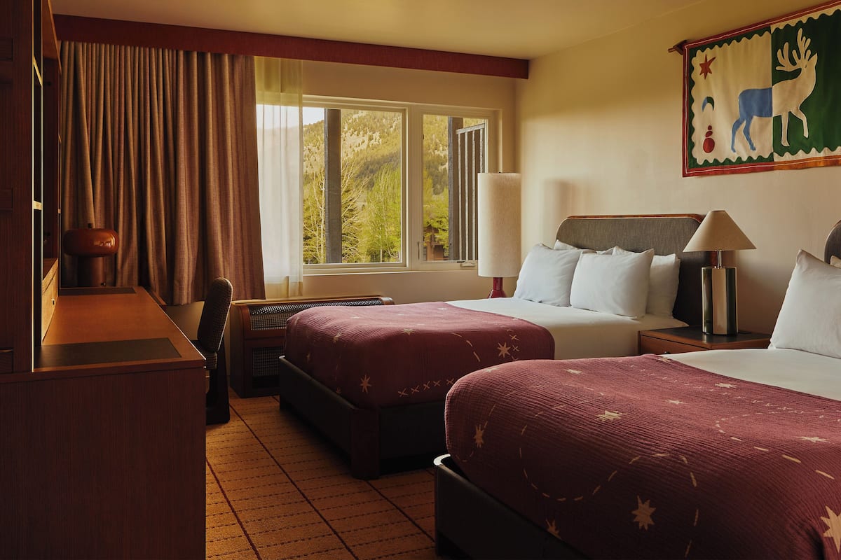 A hotel room with two beds, a desk, a chair, a lamp, and a window with a view of trees.