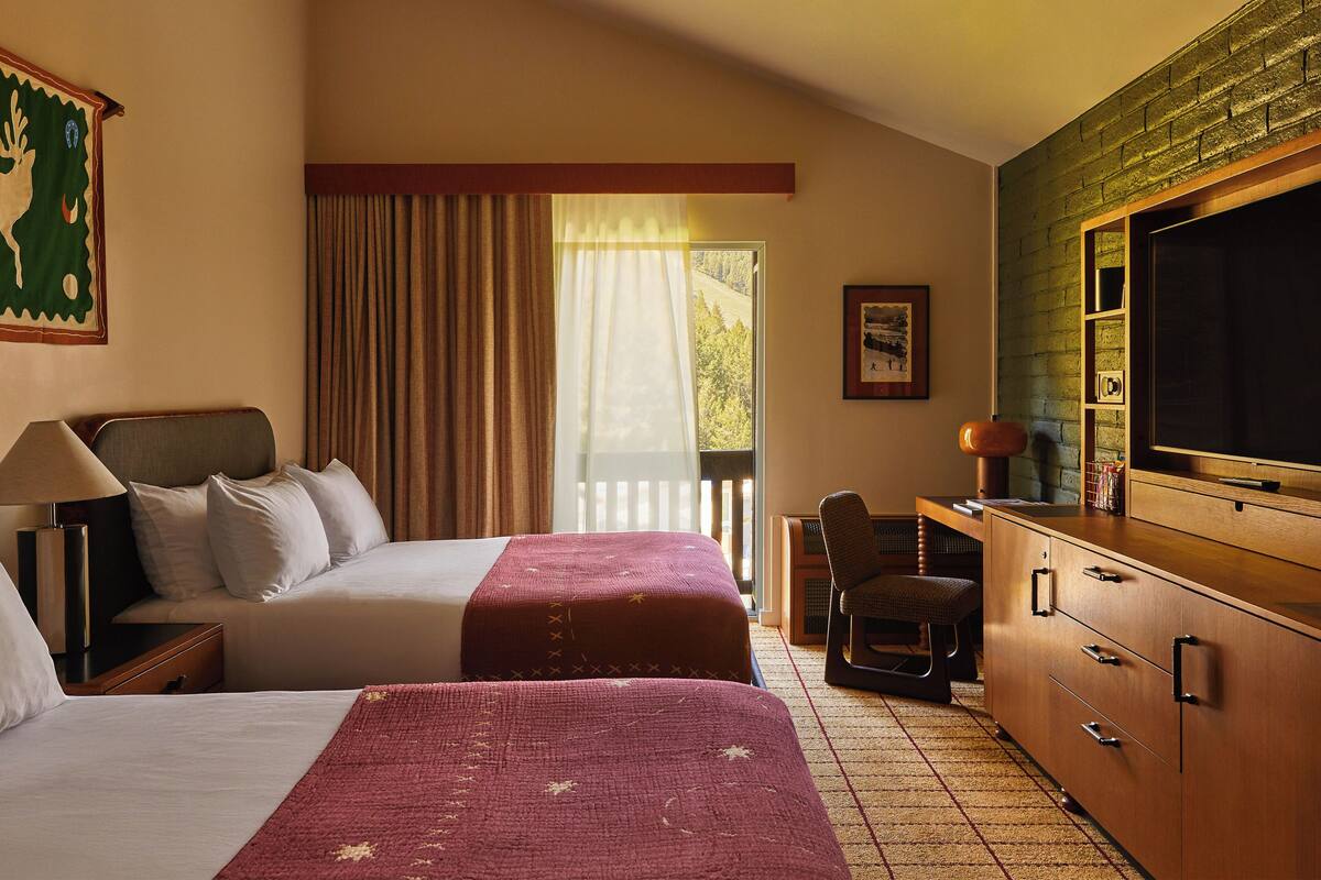 A hotel room with two beds, a desk, a chair, a TV, and a window with curtains.