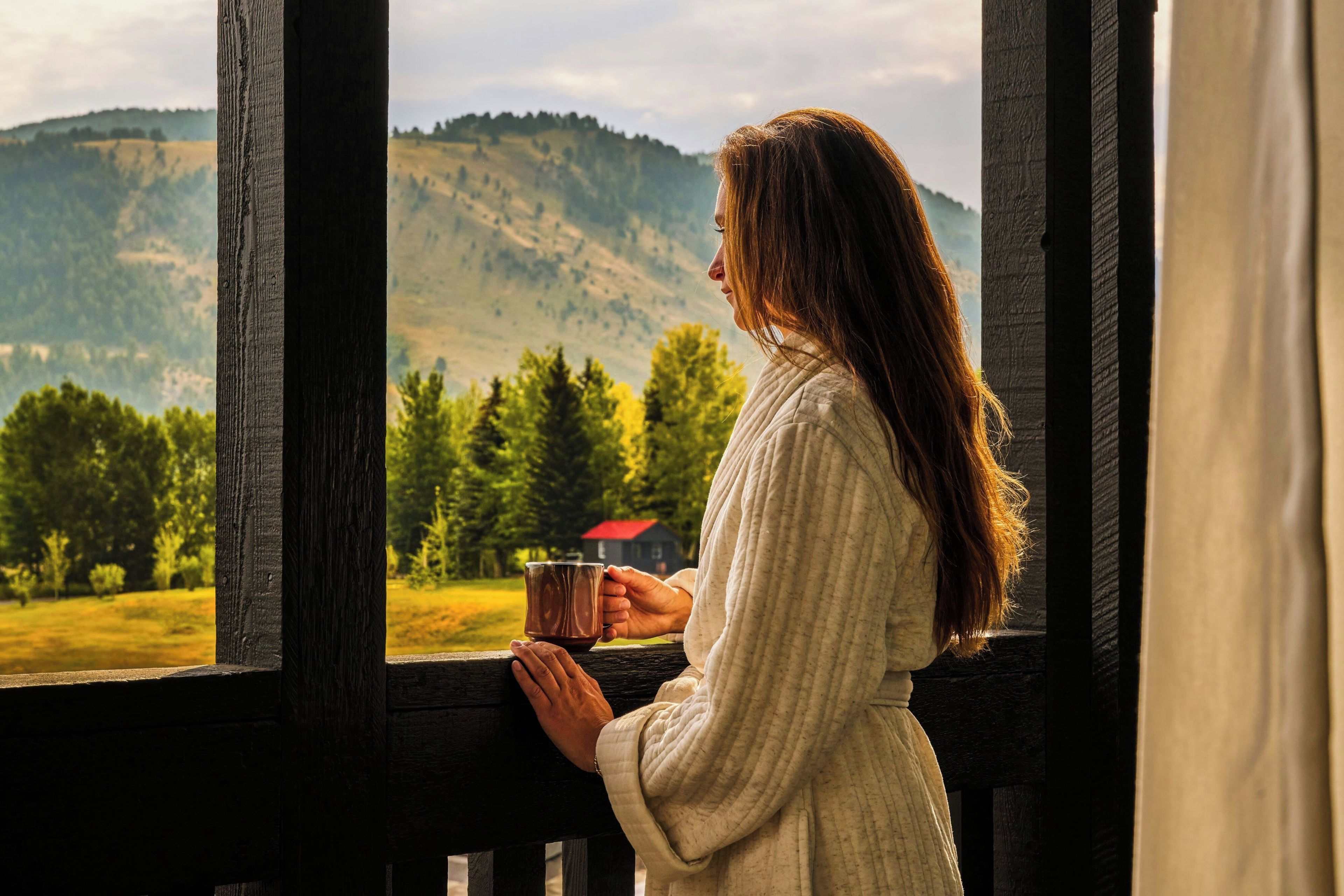 A person holding a mug, standing by a window with a view of a forest and hills.