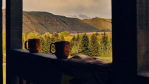 A view through a window with two mugs on a ledge, overlooking a valley with trees and distant mountains.